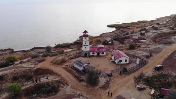 Shooting from a drone of a lighthouse at the sea against the background of hills and people walking along the road — Stock Video