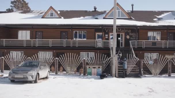 BAIKAL, IRKUTSK REGION, RUSSIA - 11 MARZO 2021: In una giornata invernale uomini e donne con borse da viaggio scendono i gradini di un autentico edificio alberghiero in legno per viaggiare in auto. Hotel villa malina — Video Stock