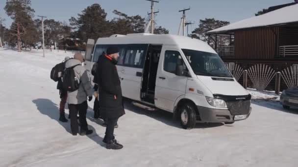 BAIKAL, IRKUTSK REGION, RUSSIE - 11 maart 2021: Een groep mensen staat bij de minibus te wachten op de tocht tegen de achtergrond van een winterlandschap en een houten gebouw — Stockvideo
