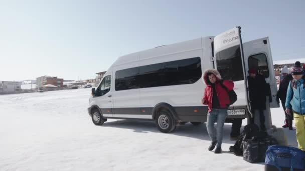 BAIKAL, IRKUTSK REGION, RUSSIA - MARCH 11, 2021: A group of young men and women stand by a white minibus on a winter day and load their travel bags before the trip — Stock Video