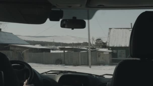Вид через лобовое стекло машины зимней дороги в деревне с деревянными домами на фоне заснеженных гор — стоковое видео