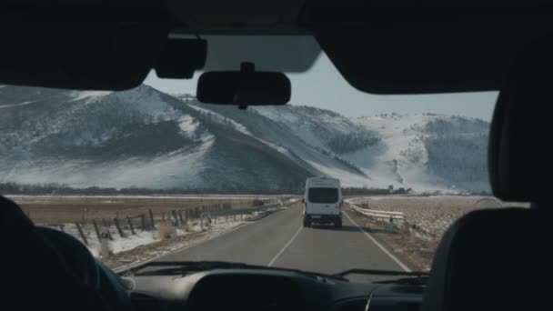 Egy autó egy kisbusz mögött vezet egy aszfaltozott úton, a hófödte hegyek hátterében. Kilátás a szélvédőn keresztül — Stock videók