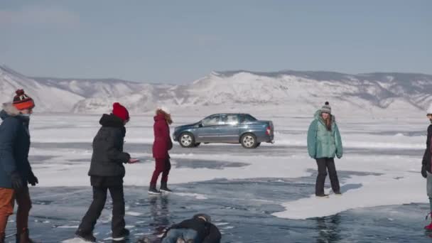 BAIKAL, IRKUTSK REGION, RUSSIE - 18 maart 2021: Een groep mensen tijdens een wintertocht loopt op het bevroren ijs van het Baikalmeer tegen de achtergrond van bergen en een passerende auto — Stockvideo