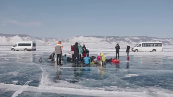 BAIKAL, REGIÓN IRKUTSK, RUSIA - 18 DE MARZO DE 2021: Un grupo de personas se levanta y se encuentra en el hielo helado del lago Baikal contra el telón de fondo de minibuses y cabos de la isla Olkhon — Vídeos de Stock
