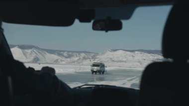 Baykal Gölü 'nün donmuş buzu üzerinde giden UAZ minibüsünün görüntüsü başka bir arabanın yolcu vagonundan dağ aralıklarıyla çevrili.