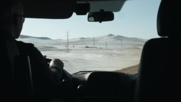 Ένας άνδρας με γυαλιά που οδηγεί ένα αυτοκίνητο οδηγεί κατά μήκος ενός χειμερινού δρόμου με φόντο τα χιονισμένα μικρά βουνά. Εσωτερική άποψη — Αρχείο Βίντεο