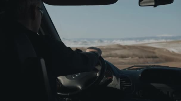 Ένας άντρας με γυαλιά οδηγεί ένα αυτοκίνητο σε ένα χειμερινό χωματόδρομο με φόντο οροσειρές κοντά στη λίμνη Baikal. Θέα από το αυτοκίνητο — Αρχείο Βίντεο