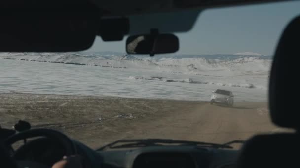 Een man rijdt een auto langs een onverharde weg langs bevroren Lake Baikal en besneeuwde bergketens. Winterreizen — Stockvideo