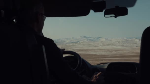 Bir kış günü araba süren bir adam, arka planda karla kaplı olmayan dağlara ve bulutlu gökyüzüne çarpar. — Stok video