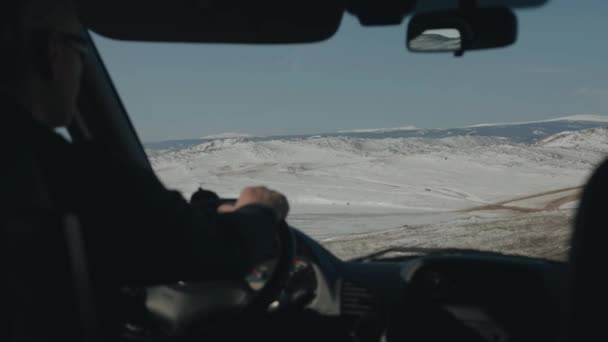 Un hombre con gafas conduce un coche en un camino de tierra de invierno contra el telón de fondo de las cadenas montañosas cerca del lago Baikal. Vista desde el coche — Vídeos de Stock