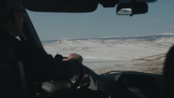 Gözlüklü bir adam, Baykal Gölü yakınlarındaki dağ sıralarının arka planına karşı kış toprak yolunda araba sürüyor. Arabadan bak. — Stok video