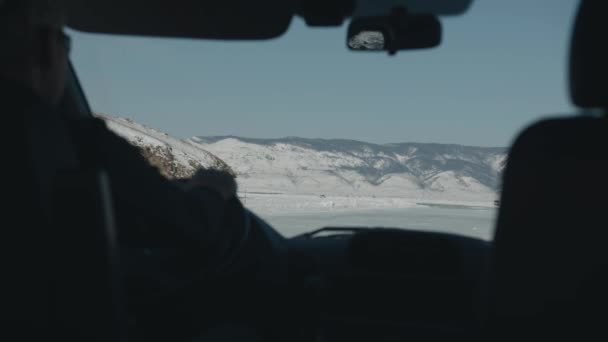Un hombre conduce un coche en un viaje de invierno por carretera a lo largo del congelado lago Baikal rodeado de montañas cubiertas de nieve. Vista interior — Vídeos de Stock
