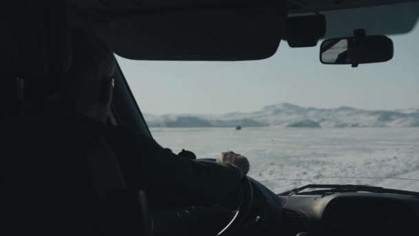 Una vista a través del parabrisas de un coche en una carretera de invierno a lo largo del lago congelado Baikal contra el telón de fondo de los cabos de la isla de Olkhon — Vídeos de Stock