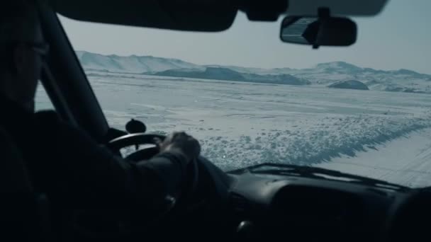 Un homme conduit une voiture sur une route d'hiver sur fond de chaînes de montagnes entourant le lac Baïkal gelé — Video