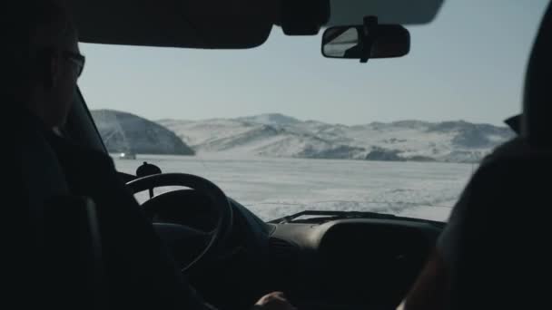 Un hombre conduce un coche en un camino de invierno contra el telón de fondo de montañas cubiertas de nieve. Vista a través del parabrisas — Vídeos de Stock