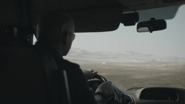 Un hombre con gafas que conduce un coche conduce a lo largo de la carretera entre la llanura y contra el fondo de las montañas en un día de invierno — Vídeos de Stock