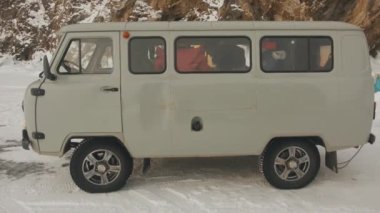 BAIKAL, IRKUTSK REGION, RUSSIA - 18 Mart 2021: Baykal Gölü 'nün donmuş buzunda dağın eteğinde bir grup turistle birlikte Minibus UAZ. Yan görünüm