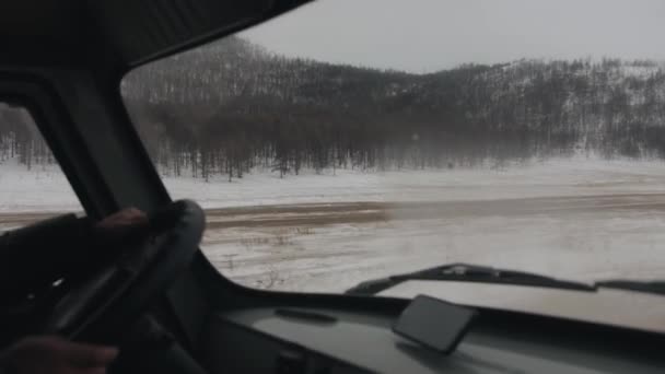 Utsikt från bilen till snötäckta berg täckta med skog och en grusväg vid foten under en vinterresa — Stockvideo
