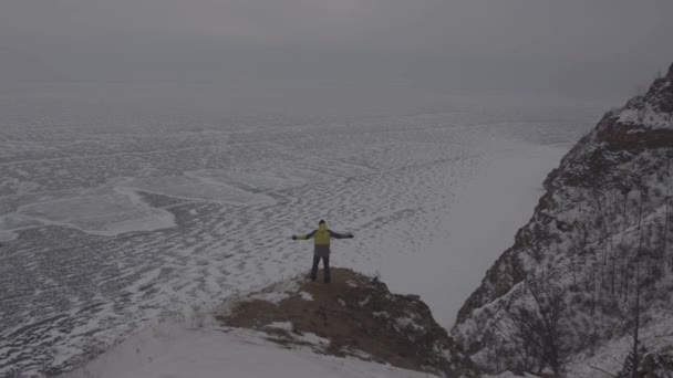 Een man staat met zijn armen uitgestrekt op een berg en bewondert het grandioze uitzicht op het Baikalmeer bedekt met ijs — Stockvideo