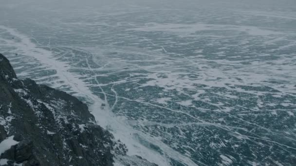 Una vista desde la montaña hasta el congelado lago Baikal con grietas en la superficie y con personas y coches a los pies — Vídeo de stock