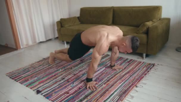 Un joven musculoso con un torso desnudo entrenando en su habitación en una alfombra y hace saltos y flexiones — Vídeo de stock