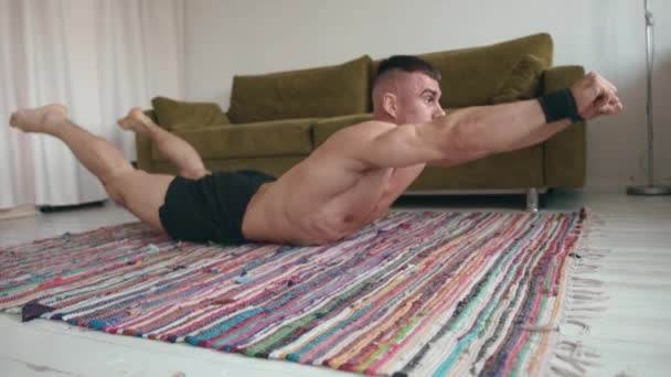 Joven culturista torso desnudo haciendo ejercicio en casa y haciendo ejercicio muscular de espalda mientras está acostado en una alfombra — Vídeo de stock