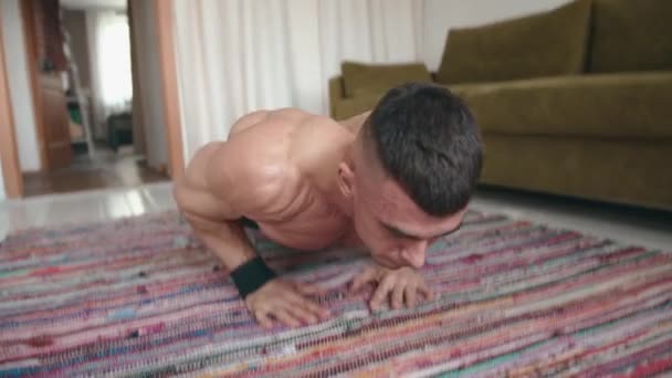 Joven hombre musculoso en pantalones cortos negros entrenando en casa y haciendo flexiones y columpios manos en tablón de ejercicios — Vídeo de stock