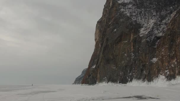 La gente camina a lo largo del hielo cubierto de nieve del lago Baikal a una enorme montaña rocosa cubierta de árboles en la orilla — Vídeos de Stock