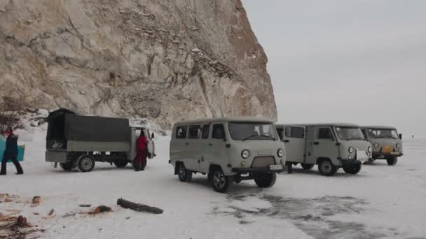 Baikal, obwód irkucki, Rosja - 18 marca 2021 r.: Minibusy UAZ stoją u podnóża skały na lodzie jeziora Bajkał i grupa ludzi udaje się do obozu namiotowego — Wideo stockowe