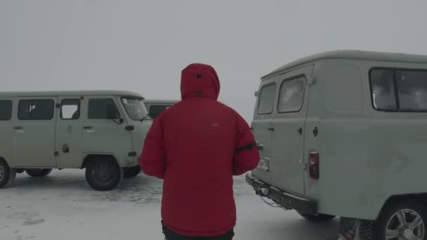 Байкал, Іркутська область, Росія - 18 березня 2021 р.: чоловік у червоній зимовій куртці з капюшоном іде між мінібусами УАЗ на замерзлому озері Байкал. Назад. Повільний рух — стокове відео