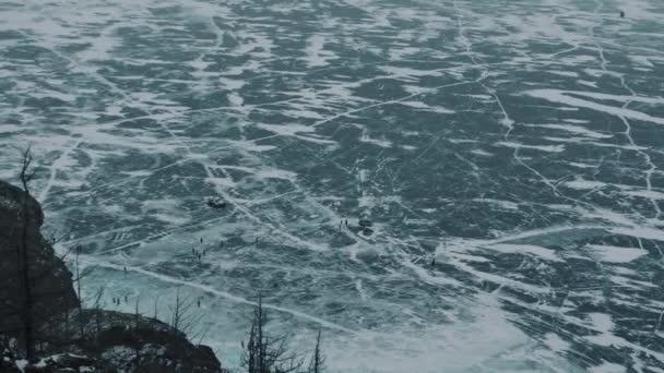 Una vista desde la montaña a los coches y la gente en el hielo congelado del lago Baikal cubierto de grietas al pie de la roca — Vídeo de stock