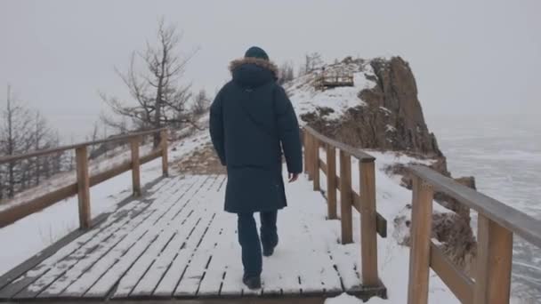 Mężczyzna w zimowej kurtce spaceruje po drewnianym moście na skale na wybrzeżu zamarzniętego jeziora Baikal. Widok z tyłu. Zwolniony ruch — Wideo stockowe