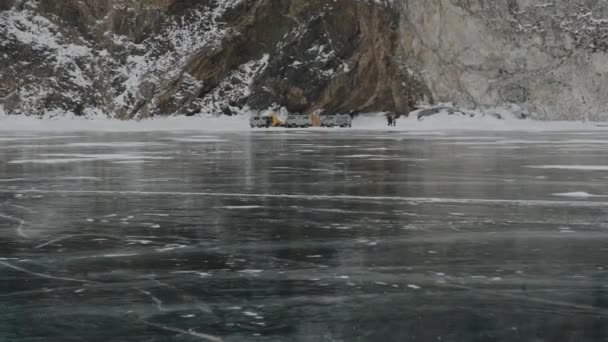 Baikal, Región de Irkutsk, Rusia - 18 de marzo de 2021: Minibuses de UAZ y un grupo de personas en un campamento de campaña en el hielo transparente del lago Baikal al pie de una montaña rocosa — Vídeo de stock