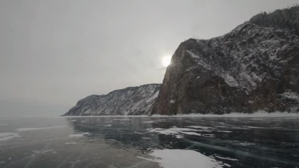 Зимний пейзаж с горами на фоне солнца, прорывающегося сквозь облака и автомобили по прозрачному льду озера Байкал — стоковое видео