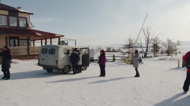 Baikal, regione di Irkutsk, Russia - 18 marzo 2021: Un gruppo di turisti si trova vicino a un minibus UAZ vicino a un hotel del villaggio e scatta foto di un paesaggio invernale con i loro telefoni cellulari. Rallentatore — Video Stock