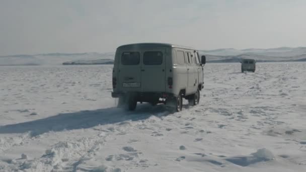 Baikal, Irkutsk Region, Russia - March 18, 2021: Δύο μικρά λεωφορεία UAZ οδηγούν σε χιονοσκεπή εξογκώματα στον πάγο της λίμνης Baikal στο φόντο των παράκτιων βράχων. Αργή κίνηση — Αρχείο Βίντεο