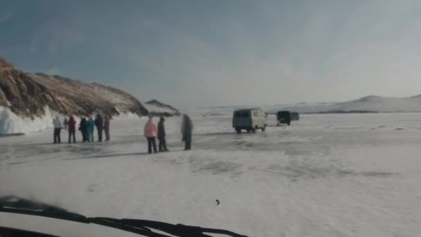 Baikal, regio Irkoetsk, Rusland - 18 maart 2021: Een weg op het ijs van het Baikalmeer met een kolom UAZ-minibussen en een groep mensen die langs de bevroren kust lopen — Stockvideo