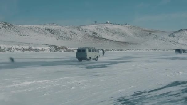 Minibuses UAZ están conduciendo a lo largo de la carretera en el lago congelado Baikal y grupos de turistas caminando sobre el hielo cerca de las rocas costeras — Vídeos de Stock