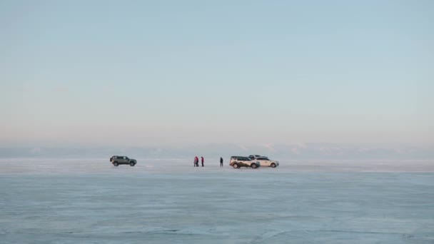 Turyści chodzą do samochodów na lodzie zamarzniętego jeziora Baikal na tle ośnieżonych skał na brzegu — Wideo stockowe
