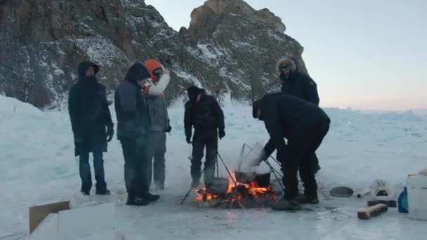 Baikal, Región de Irkutsk, Rusia - 18 de marzo de 2021: Los hombres cocinan comida en macetas sobre un fuego en un campamento de tiendas de campaña en el hielo del lago Baikal, cerca de las costas rocosas de la isla de Olkhon — Vídeo de stock