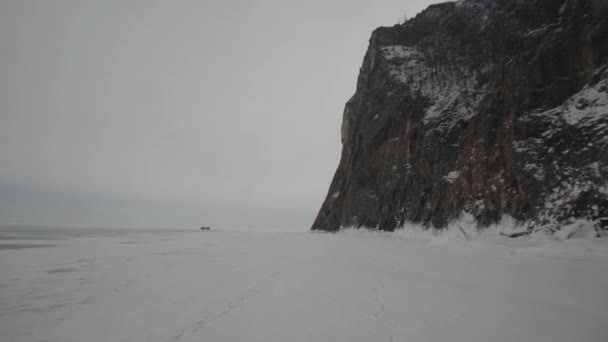 Una hermosa vista de una montaña rocosa sobre el fondo de un cielo gris y coches de pie sobre el hielo del lago Baikal — Vídeos de Stock