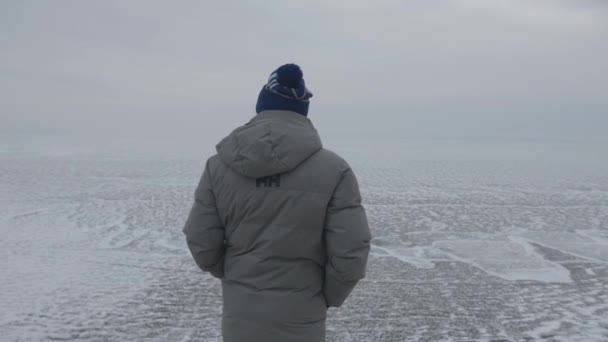 Baikal, Irkutsk Region, Ryssland - 18 mars 2021: En man står på toppen av ett berg och tittar på den istäckta sjön Baikal. Begreppet frihet ett med sig själv — Stockvideo