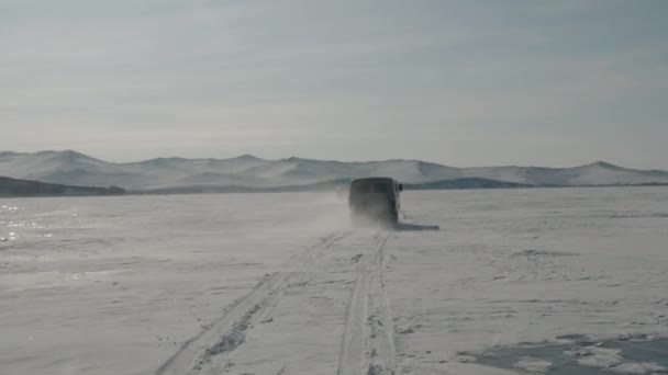 Dos minibuses UAZ viajan sobre protuberancias cubiertas de nieve en el hielo del lago Baikal contra el telón de fondo de rocas costeras. Movimiento lento — Vídeo de stock