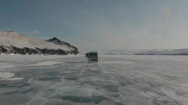 Sarake pikkubusseja UAZ suurella nopeudella ratsastaa läpinäkyvällä jäällä järven Baikal ohi kallioiset vuoret rannikolla — kuvapankkivideo