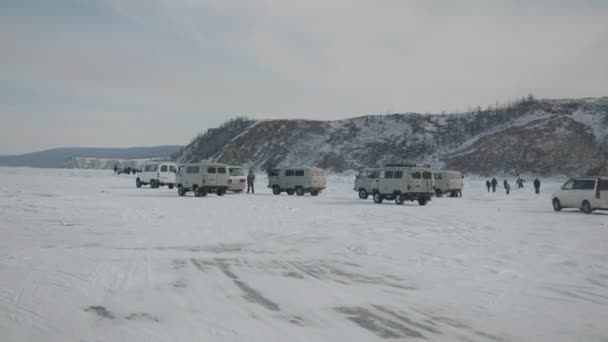 Baikal, Irkutsk Region, Russia - March 18, 2021: Θέα από το αυτοκίνητο σε αυτοκινητοπομπή λεωφορείων και τουριστών UAZ στους πρόποδες ενός γκρεμού στην ακτή της παγωμένης λίμνης Baikal — Αρχείο Βίντεο