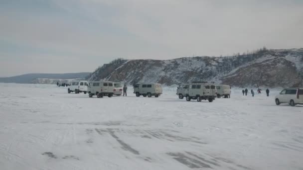 Байкал, Иркутская область, Россия - 18 марта 2021 года: Вид с автомобиля на колонну микроавтобусов УАЗ и туристов у подножия скалы на берегу замёрзшего озера Байкал — стоковое видео