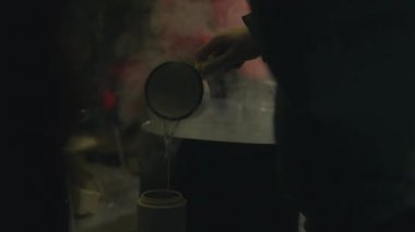 Elinde demir bardakla bir elin yakın çekimi büyük bir tencereden termosa kaynar su döküyor. Karanlık bir odada, buhar bulutlarının arasında.