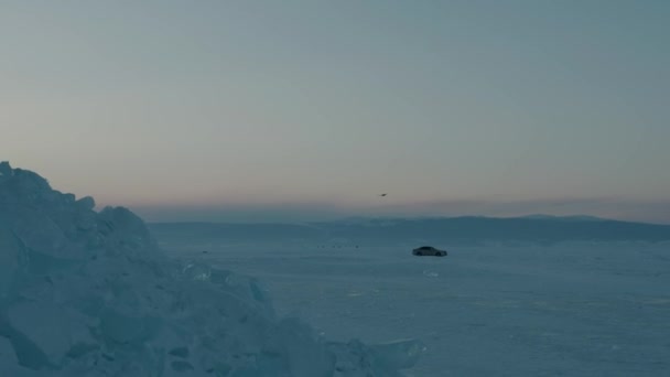 Птица пролетает над огромными прозрачными блоками голубого льда и туристами, идущими по замерзшему озеру Байкал на закате. Птица летит — стоковое видео