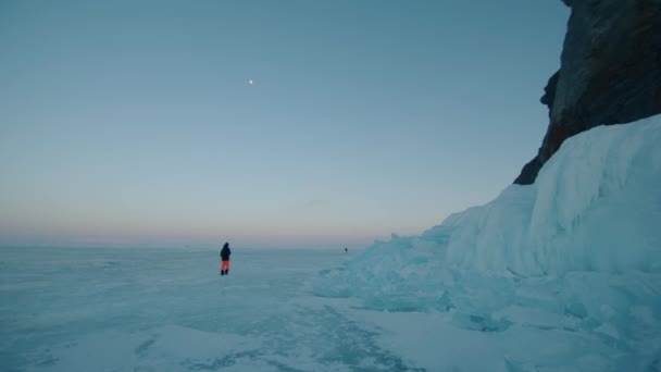 På kvällen går en man längs den frusna sjön Baikal förbi enorma blåa isblock vid foten av en klippa. Långsamma rörelser — Stockvideo