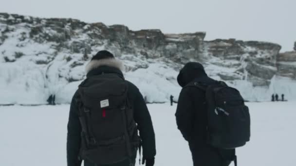 Baikal, Irkutsk Region, Russia - March 18, 2021: Δύο άνδρες πηγαίνουν κατά μήκος του παγωμένου πάγου της λίμνης Baikal στα βράχια που καλύπτονται με παγωμένες πιτσιλιές. Πίσω όψη. Αργή κίνηση — Αρχείο Βίντεο